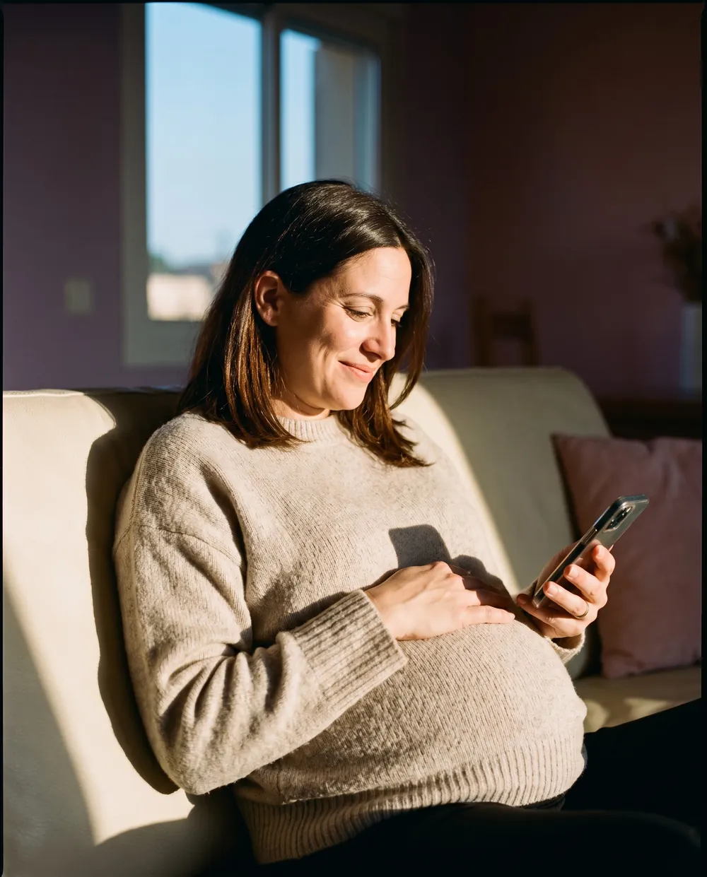 A pregnant woman smiling softly on her sofa, one hand on her belly, writing in her diary on her phone.